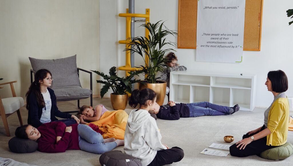 Kinder im Alter von etwa 10-12 Jahren sitzen und liegen auf dem Teppichboden und hören einer Geschichte einer erwachsenen Frau zu. Es herrscht eine gemütliche Atmosphäre.