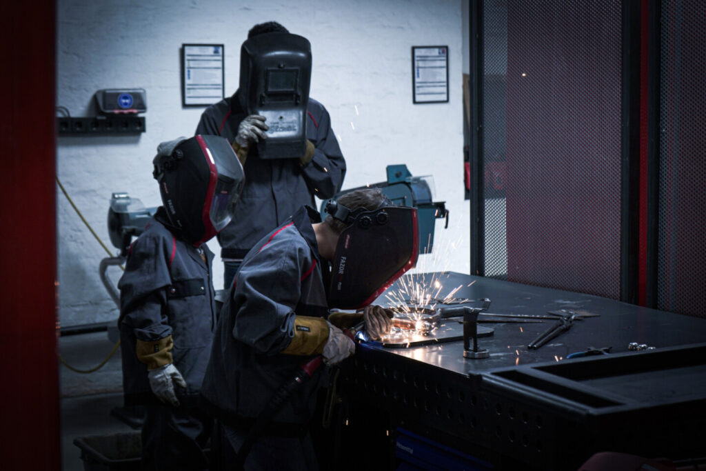 Drei Personen haben Schweißschutz Helme auf und betätigen sich beim Schweißen in der Grand Garage Werkstatt.